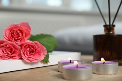 Photo of Burning candles, rose flowers, book and reed diffuser on wooden table indoors