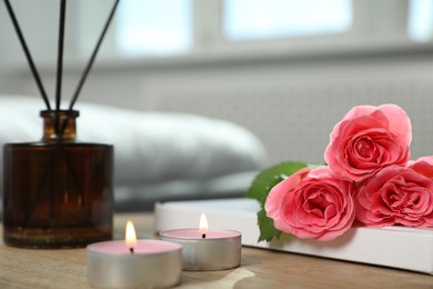 Photo of Burning candles, rose flowers, book and reed diffuser on wooden table indoors