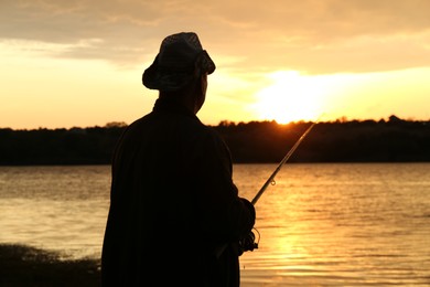 Fisherman with rod fishing near lake at sunset, back view