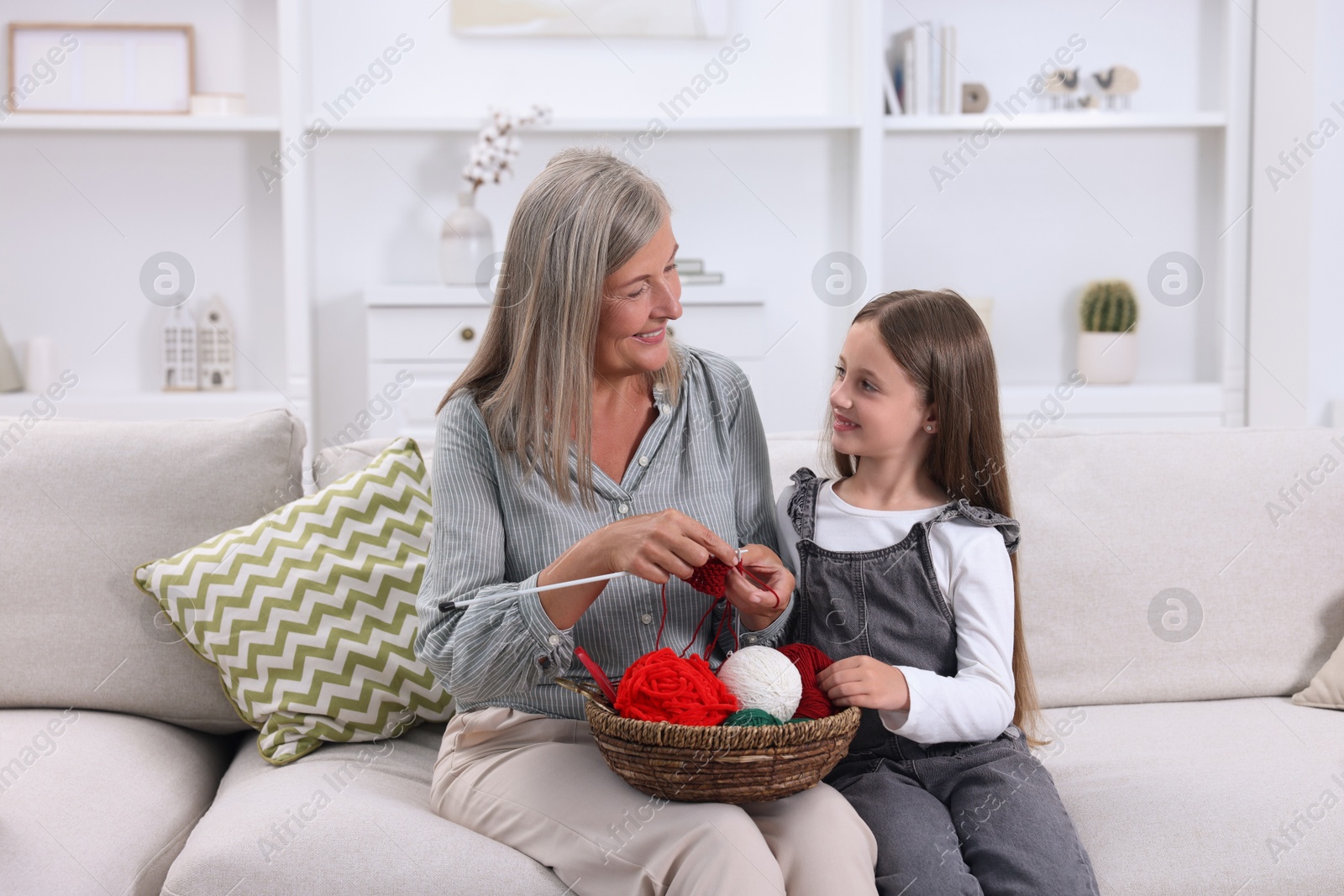 Photo of Smiling grandmother teaching her granddaughter to knit on sofa at home