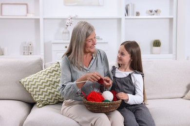 Photo of Smiling grandmother teaching her granddaughter to knit on sofa at home