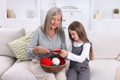 Smiling grandmother teaching her granddaughter to knit on sofa at home