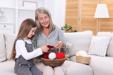 Smiling grandmother teaching her granddaughter to knit on sofa at home