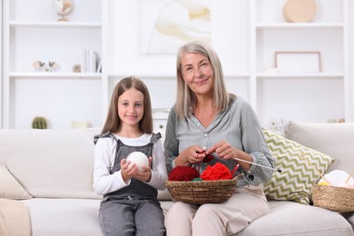 Happy grandmother with granddaughter holding knitting needles and yarn on sofa at home