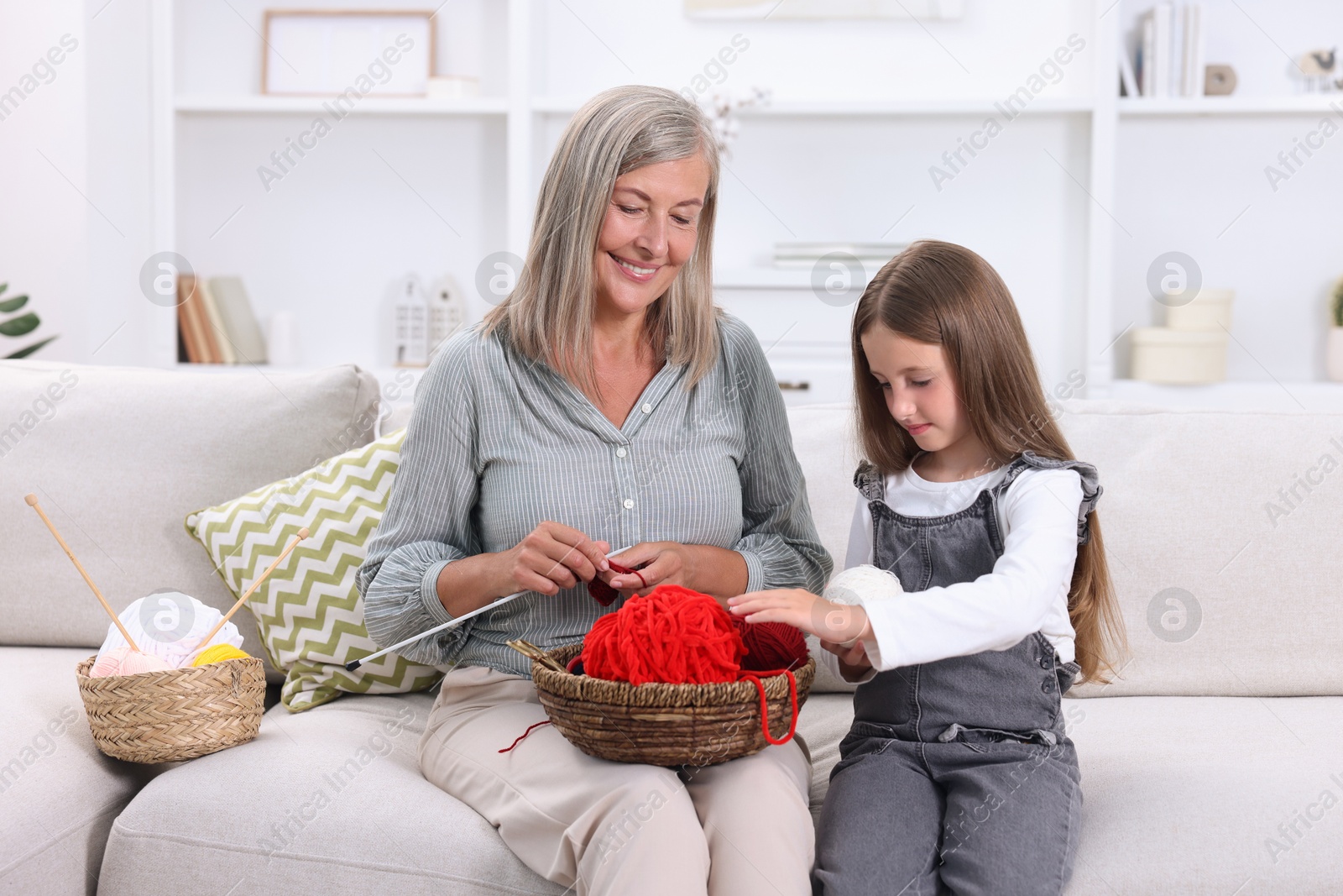 Photo of Smiling grandmother teaching her granddaughter to knit on sofa at home