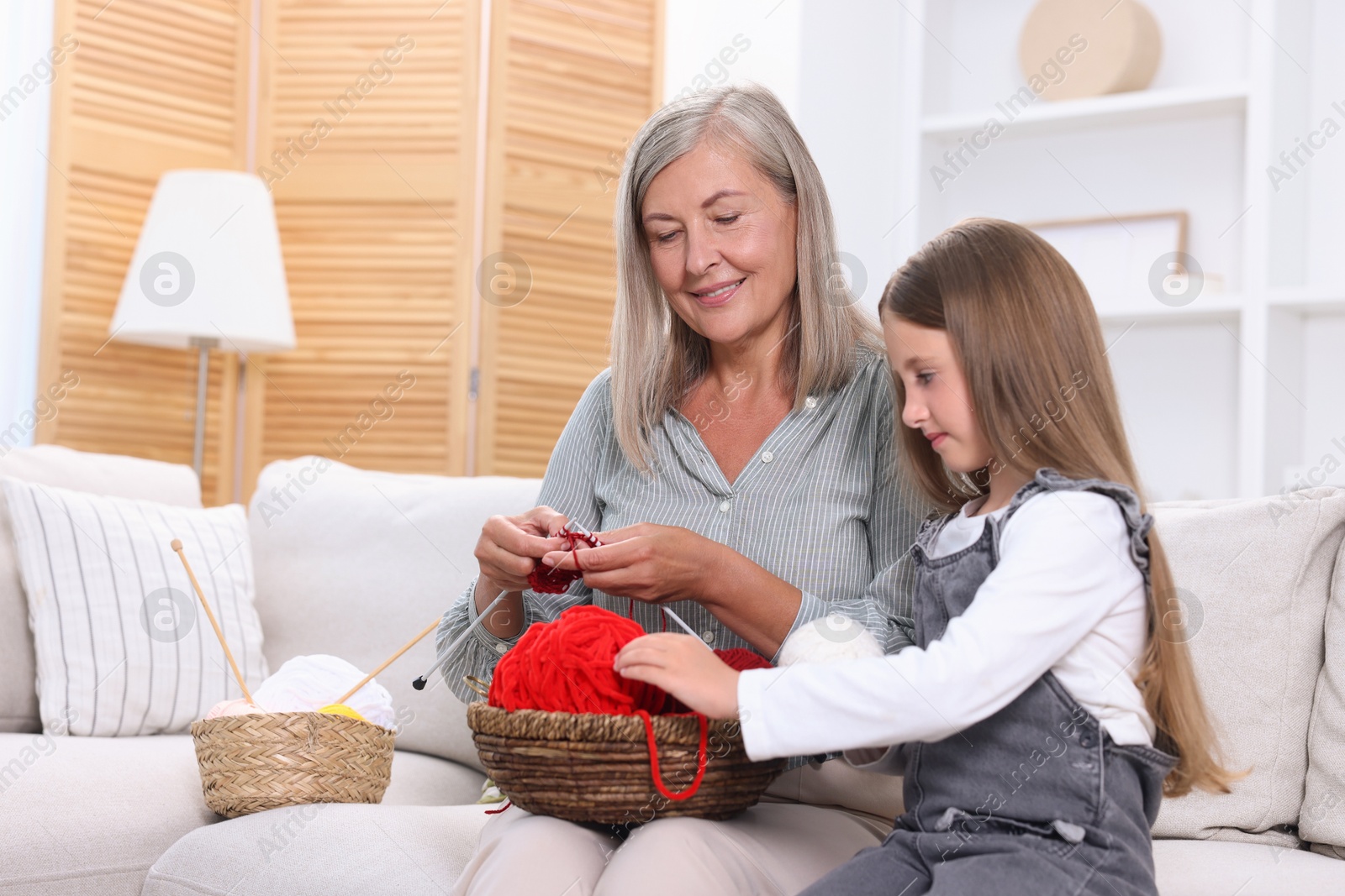 Photo of Smiling grandmother teaching her granddaughter to knit on sofa at home