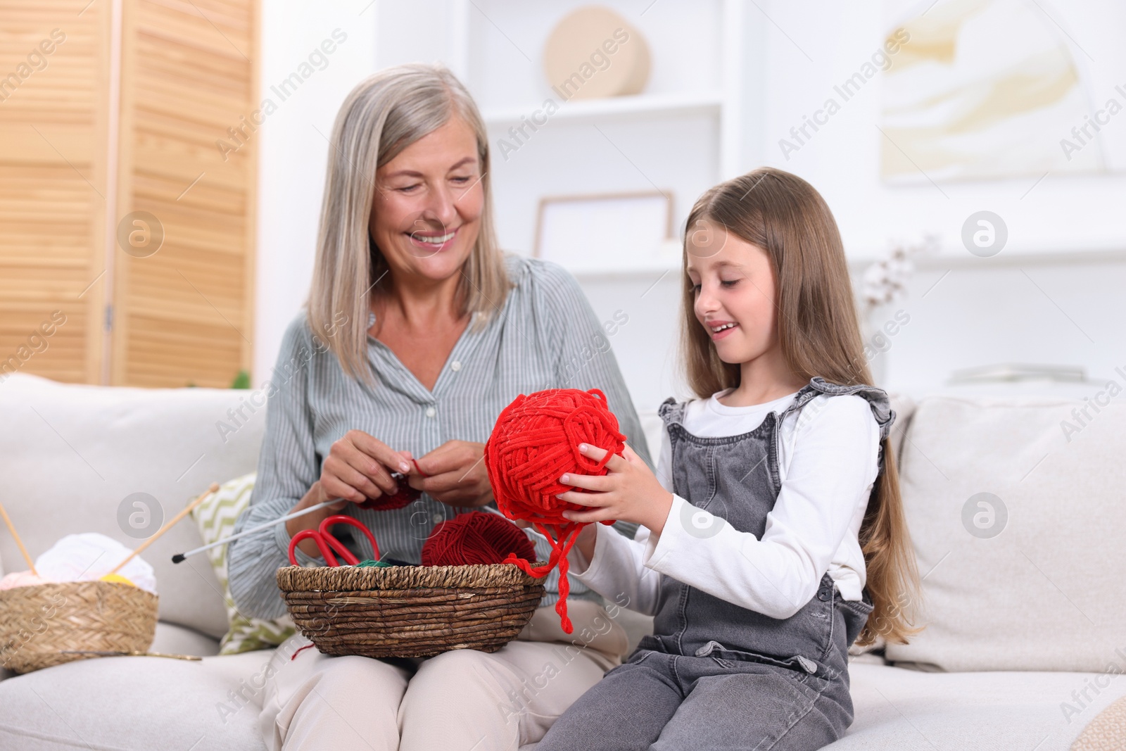 Photo of Smiling grandmother teaching her granddaughter to knit on sofa at home