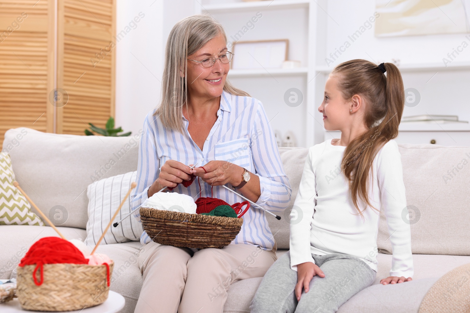 Photo of Smiling grandmother teaching her granddaughter to knit on sofa at home