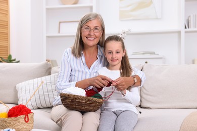 Smiling grandmother teaching her granddaughter to knit on sofa at home