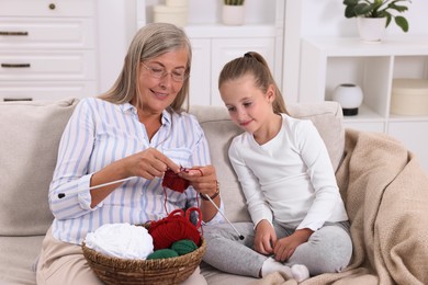 Smiling grandmother teaching her granddaughter to knit on sofa at home