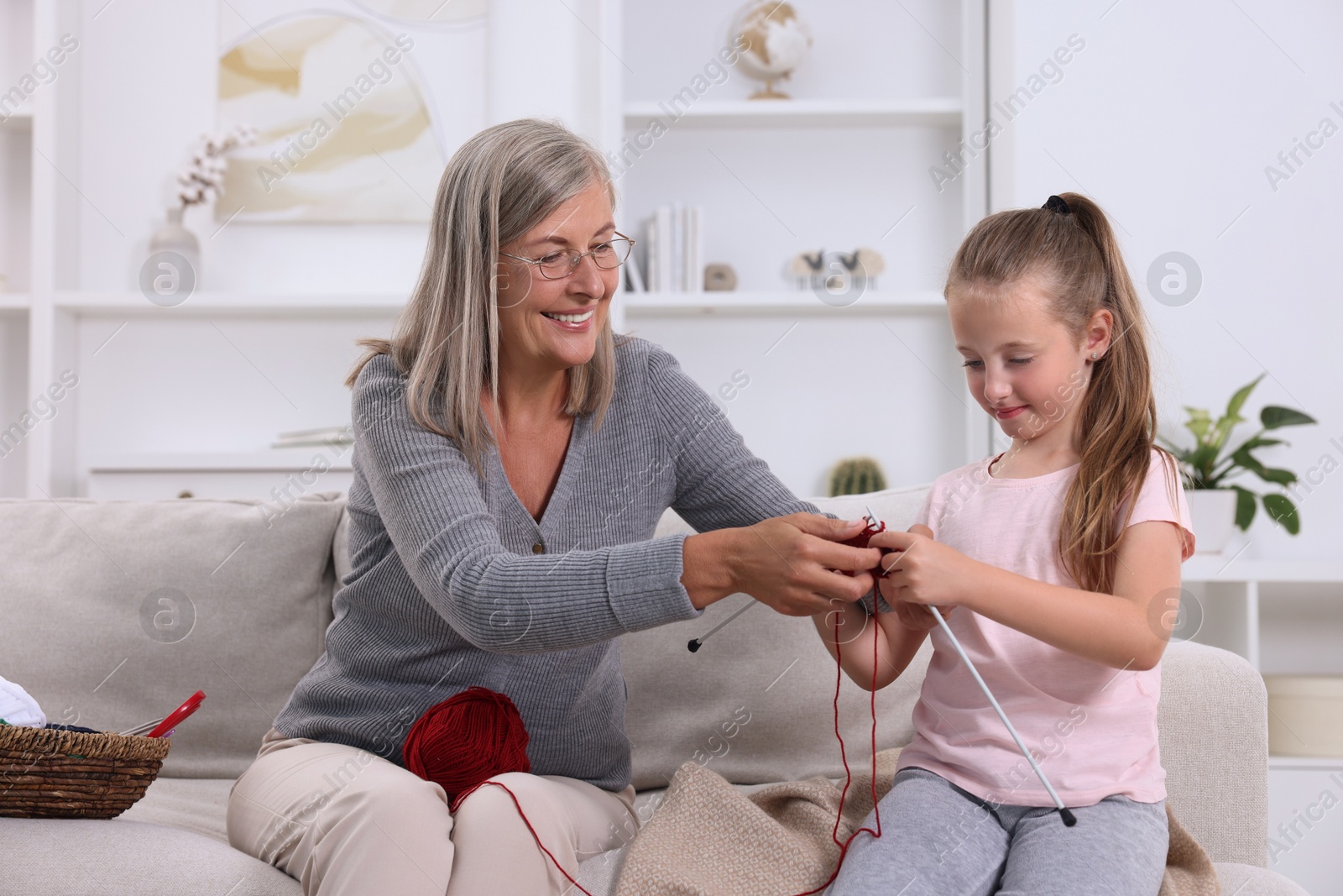 Photo of Smiling grandmother teaching her granddaughter to knit on sofa at home