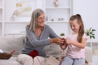 Smiling grandmother teaching her granddaughter to knit on sofa at home