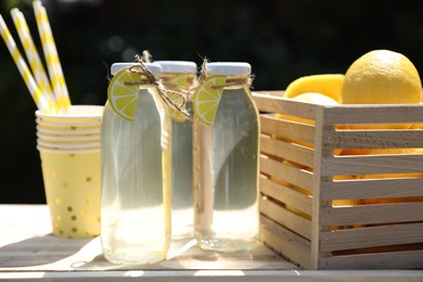 Refreshing lemonade in bottles, fresh fruits, paper cups and straws on wooden table outdoors