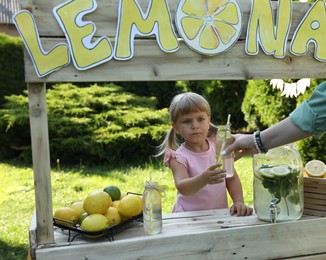 Photo of Cute little girl selling natural lemonade to woman in park