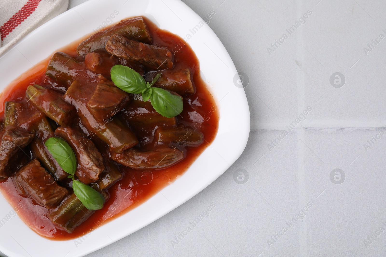 Photo of Tasty stew with okra, tomato sauce and basil on white tiled table, top view. Space for text