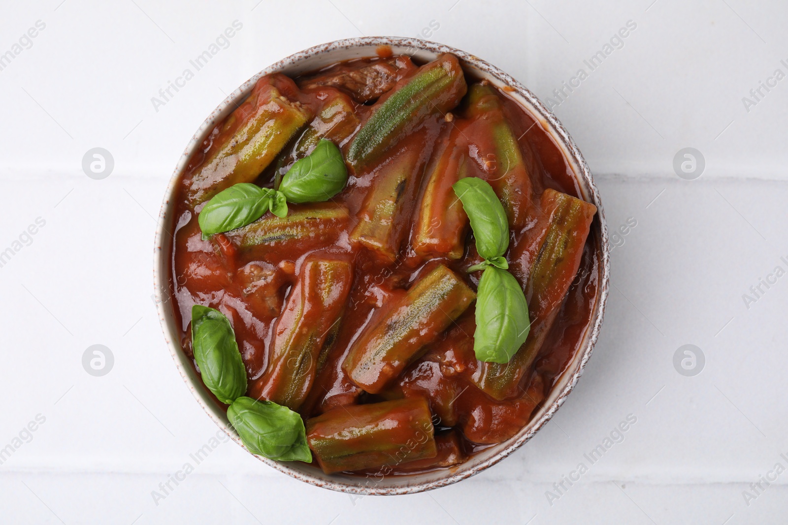 Photo of Tasty stew with okra, tomato sauce and basil on white tiled table, top view