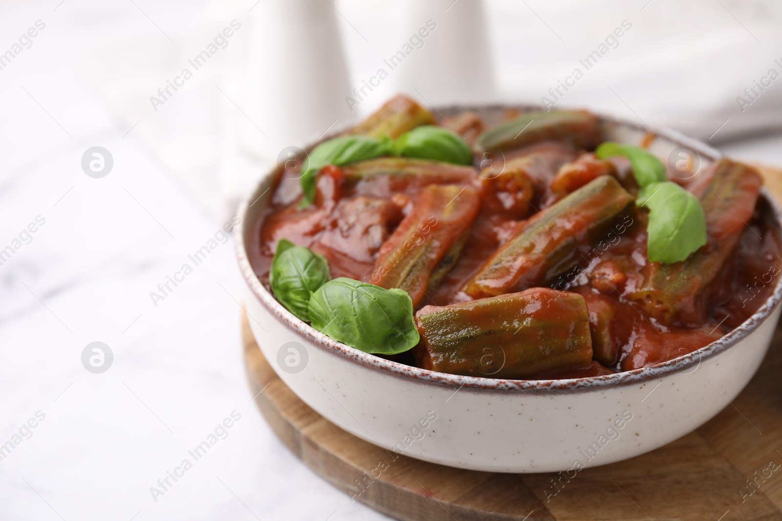 Photo of Tasty stew with okra, tomato sauce and basil on white marble table, closeup. Space for text