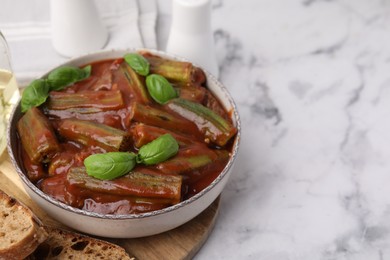 Tasty stew with okra, tomato sauce and basil on white marble table, closeup. Space for text