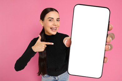 Happy woman pointing at mobile phone with blank screen on pink background. Mockup for design