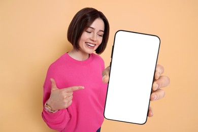 Happy woman pointing at mobile phone with blank screen on dark beige background. Mockup for design