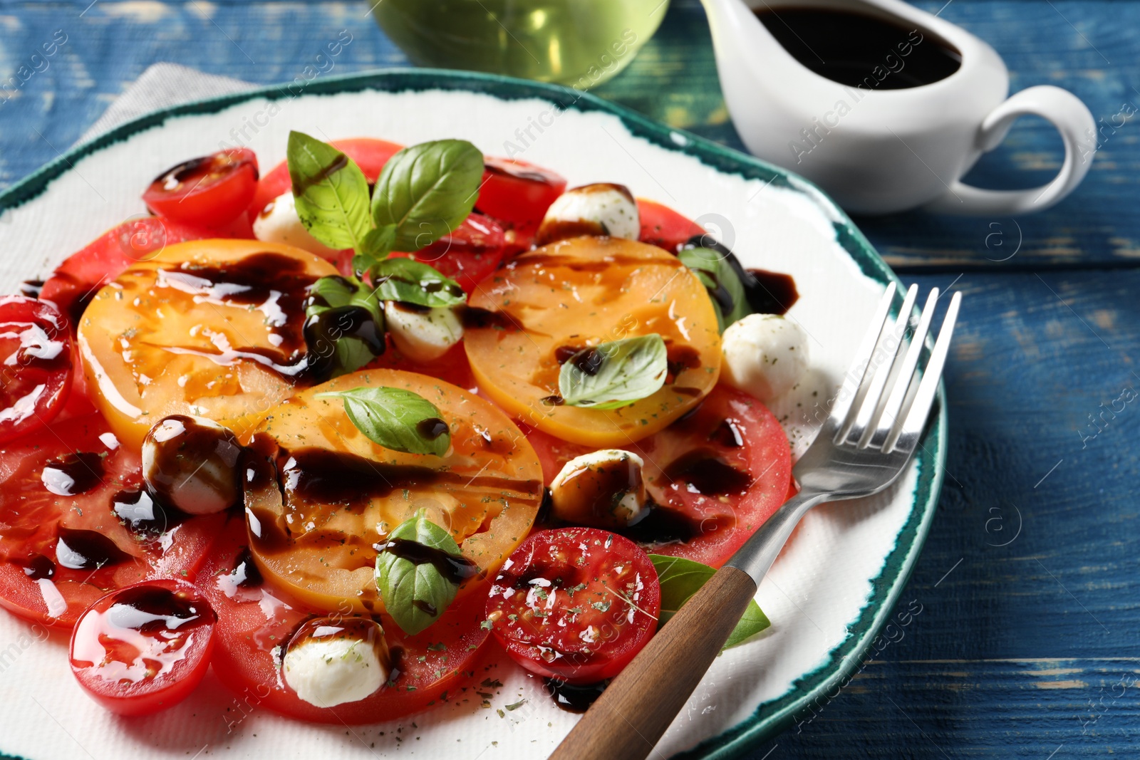 Photo of Tasty salad with balsamic vinegar, mozzarella and fork on blue wooden table, closeup