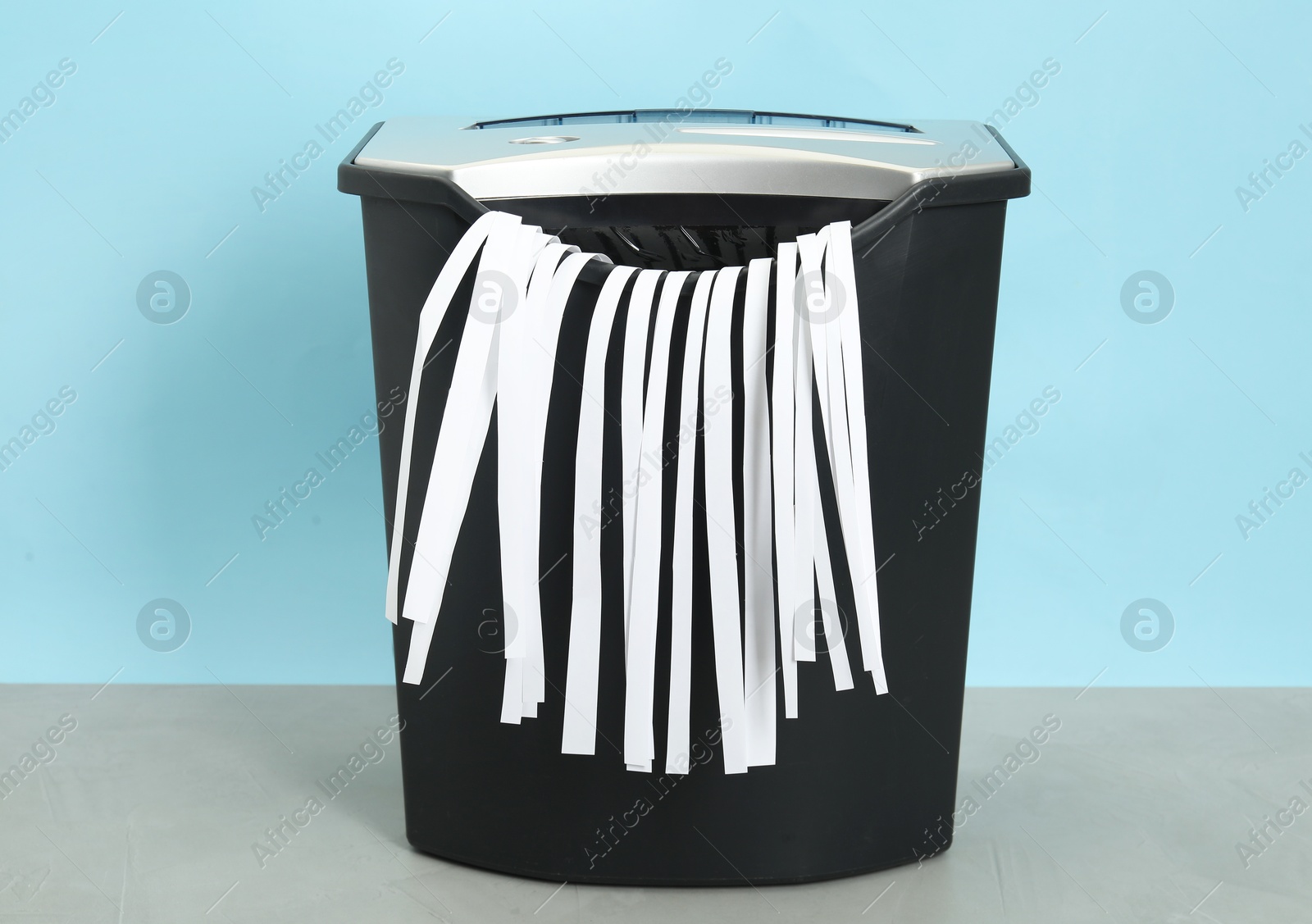 Photo of Shredder with paper strips on grey table against light blue background