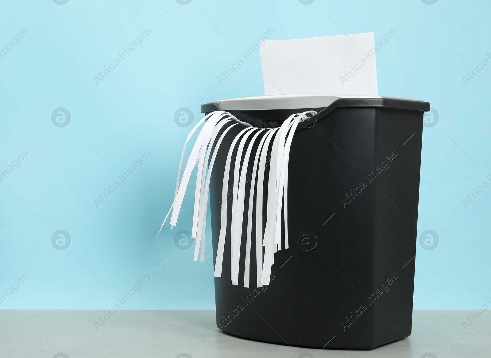 Photo of Destroying sheet of paper with shredder at grey table against light blue background, space for text