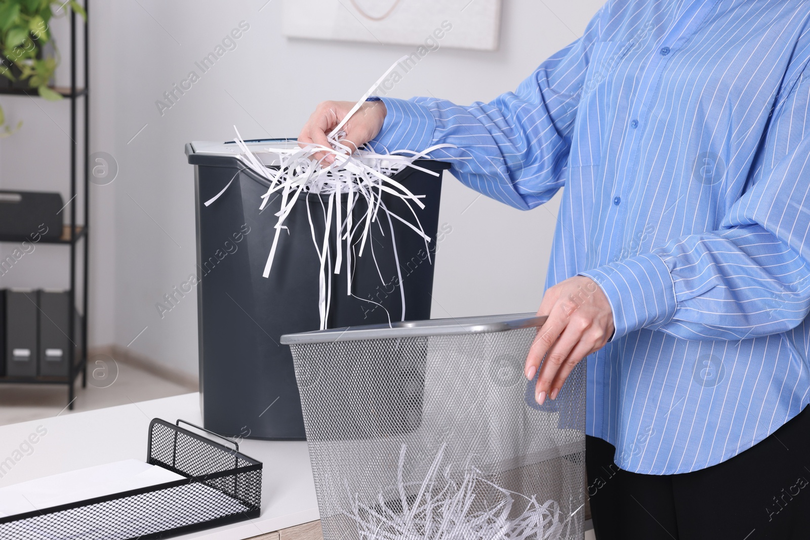 Photo of Woman putting shredded paper strips into trash bin at white table in office, closeup