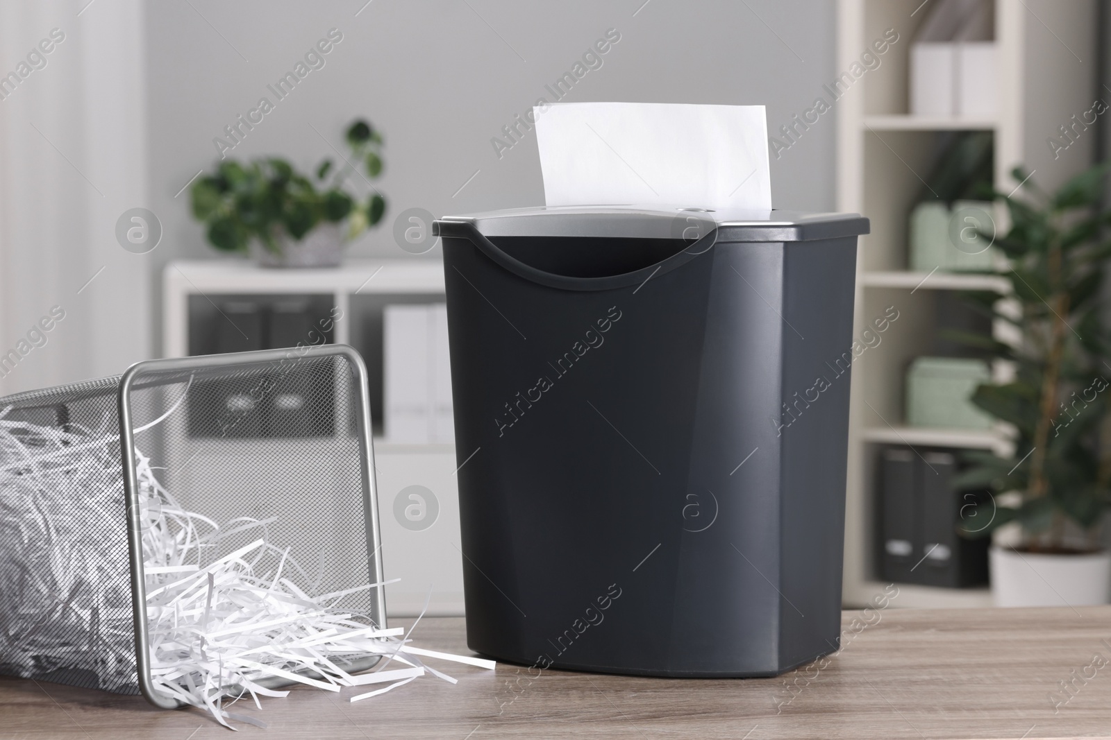 Photo of Destroying sheets with shredder and paper strips in metal bin on wooden table in office