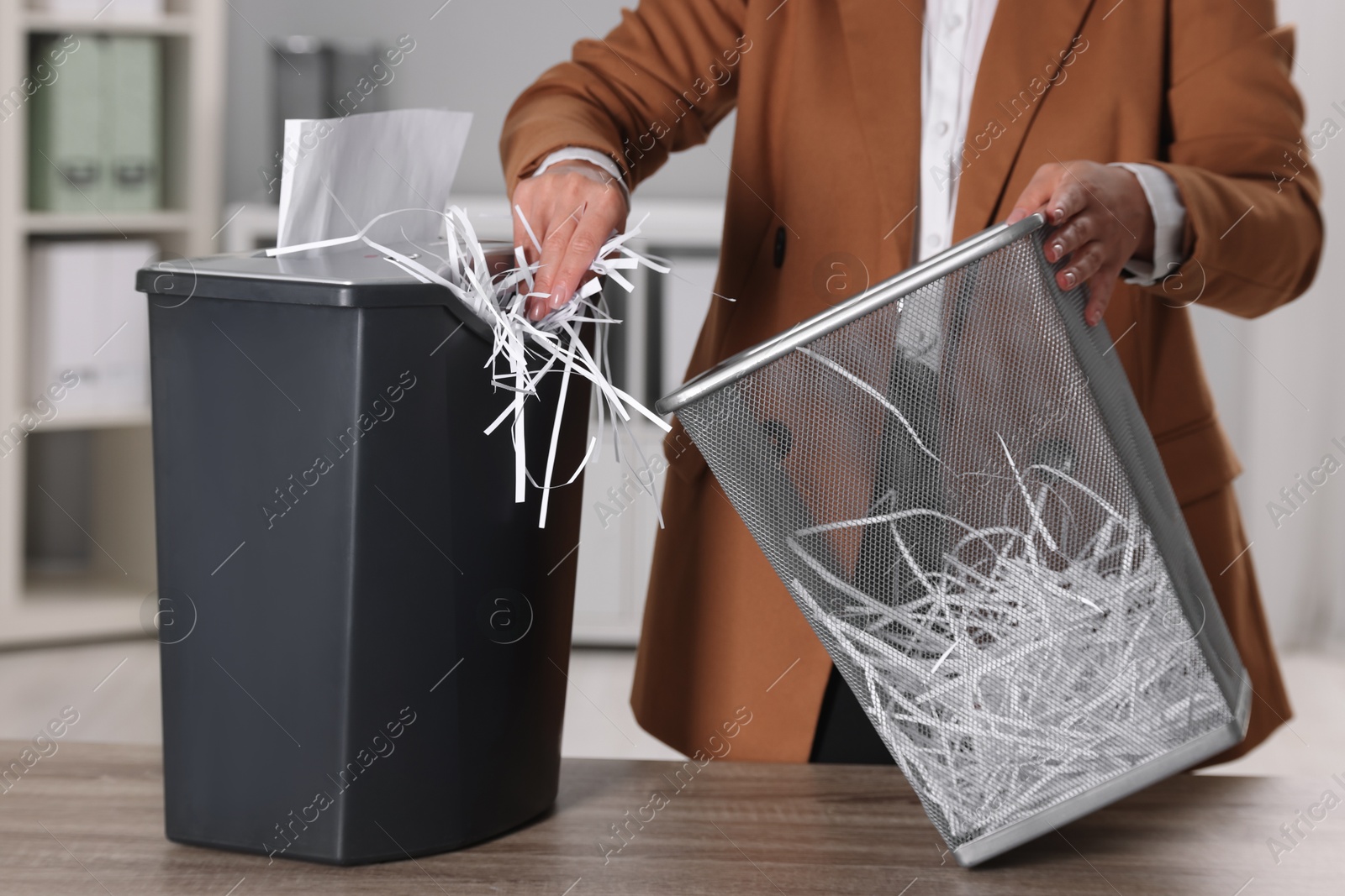 Photo of Woman putting shredded paper strips into trash bin at wooden table in office, closeup