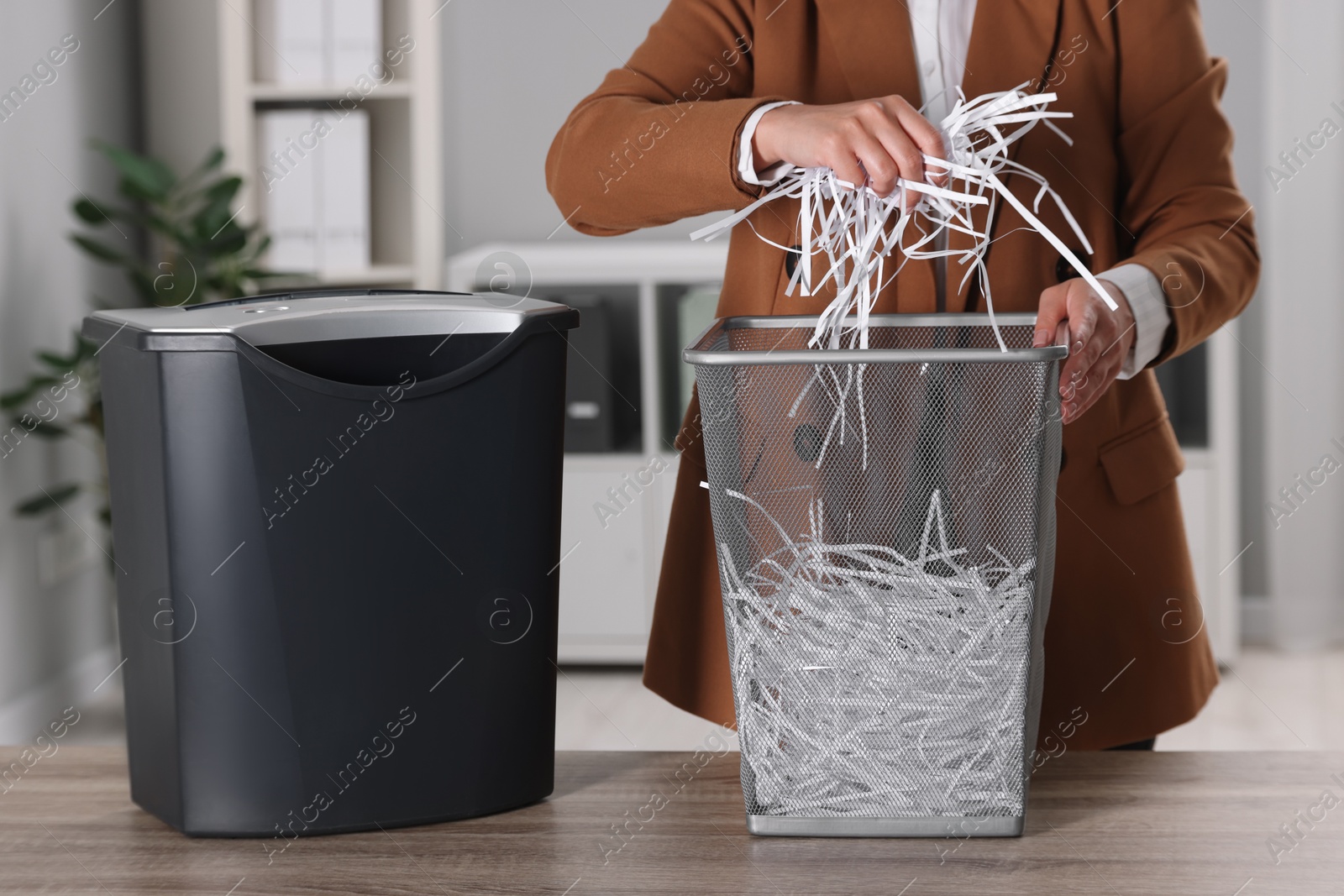 Photo of Woman putting shredded paper strips into trash bin at wooden table in office, closeup