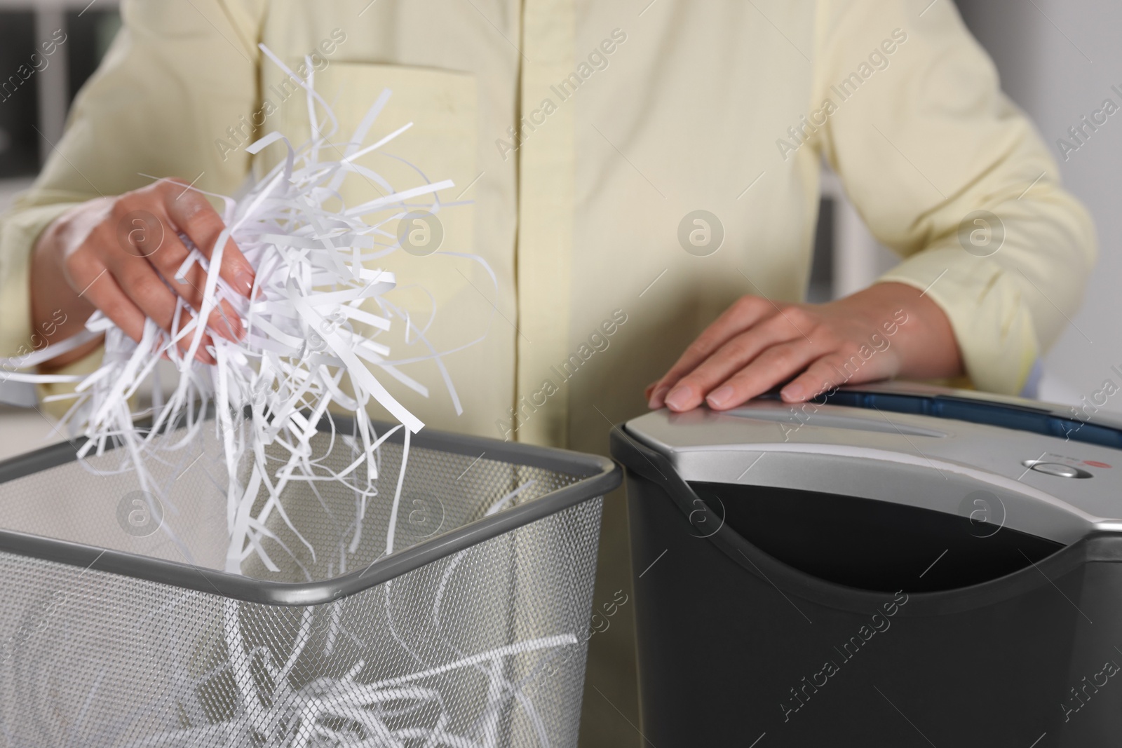 Photo of Woman putting shredded paper strips into trash bin indoors, closeup