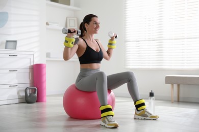 Beautiful woman with ankle weights and dumbbells training on fitness ball indoors