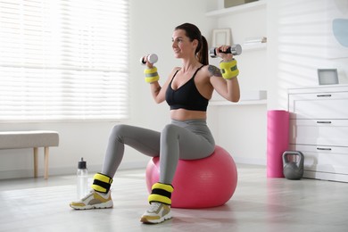 Beautiful woman with ankle weights and dumbbells training on fitness ball indoors