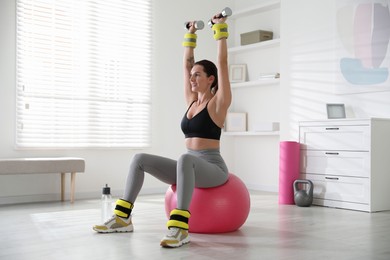 Photo of Beautiful woman with ankle weights and dumbbells training on fitness ball indoors