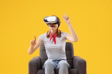 Happy woman with virtual reality headset sitting in armchair on yellow background