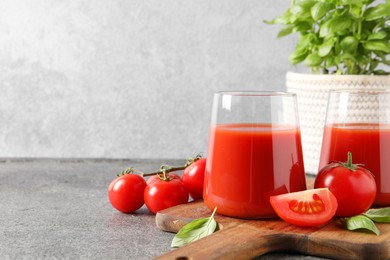 Photo of Tasty tomato juice in glasses, basil and fresh vegetables on grey table, space for text