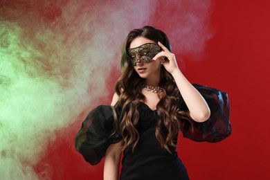 Photo of Beautiful woman wearing carnival mask on red background in color lights and smoke