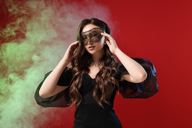 Beautiful woman wearing carnival mask on red background in color lights and smoke