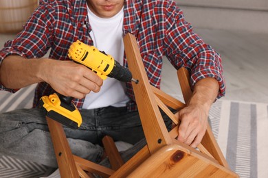 Photo of Man repairing wooden stool with electric screwdriver indoors, closeup