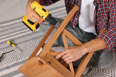 Photo of Man repairing wooden stool with electric screwdriver indoors, closeup