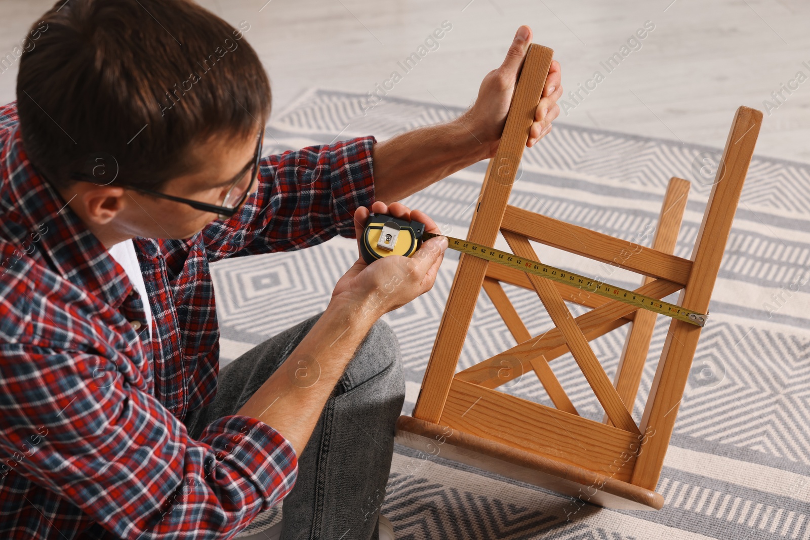 Photo of Man using tape measure while repairing wooden stool indoors