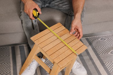 Photo of Man using tape measure while repairing wooden stool indoors, closeup