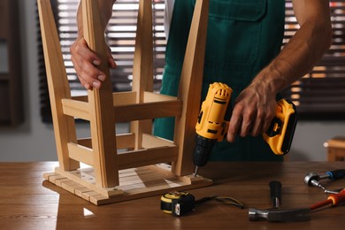 Photo of Man repairing wooden stool with electric screwdriver indoors, closeup