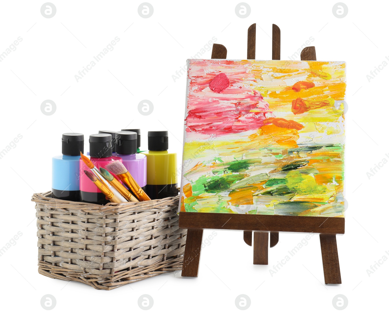 Photo of Acrylic paints of different colors with brushes in wicker basket and abstract picture on small easel against white background