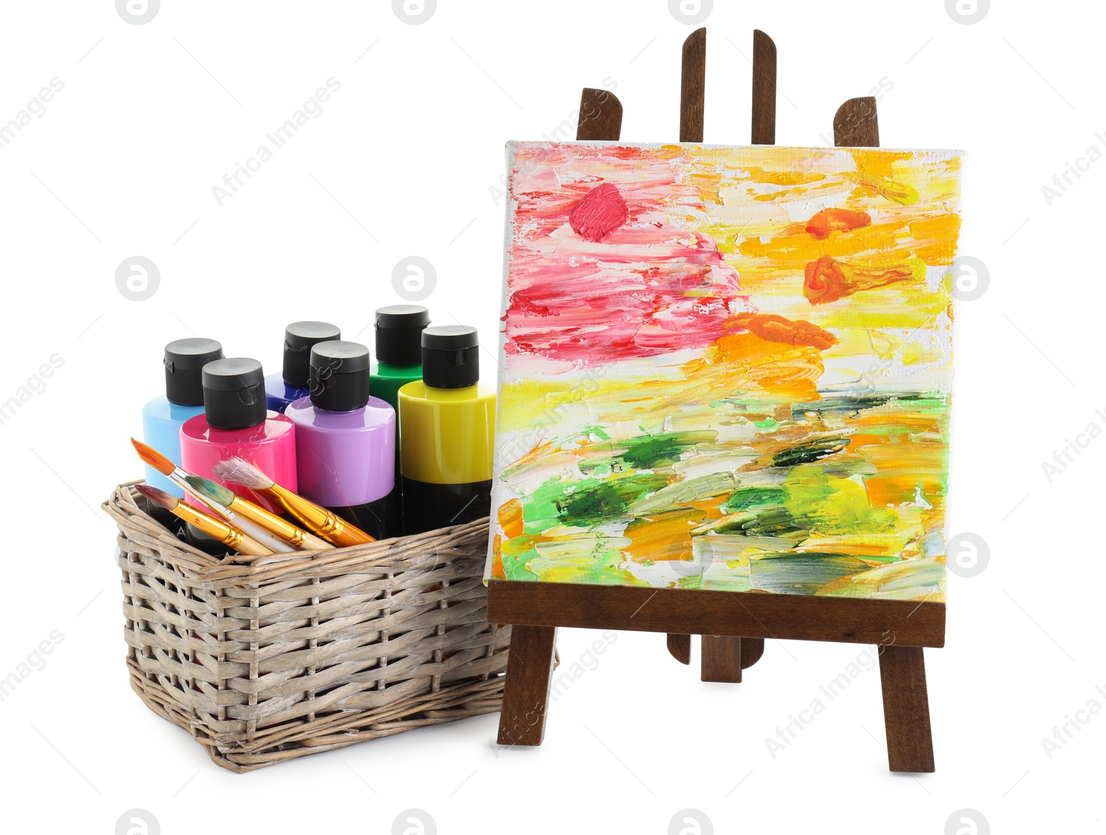 Photo of Acrylic paints of different colors with brushes in wicker basket and abstract picture on small easel against white background