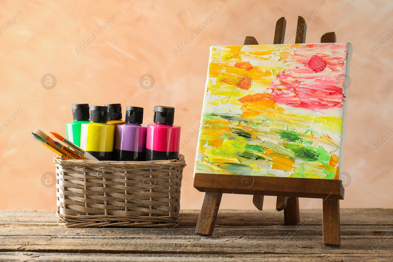 Photo of Acrylic paints of different colors with brushes in wicker basket and abstract picture on wooden table