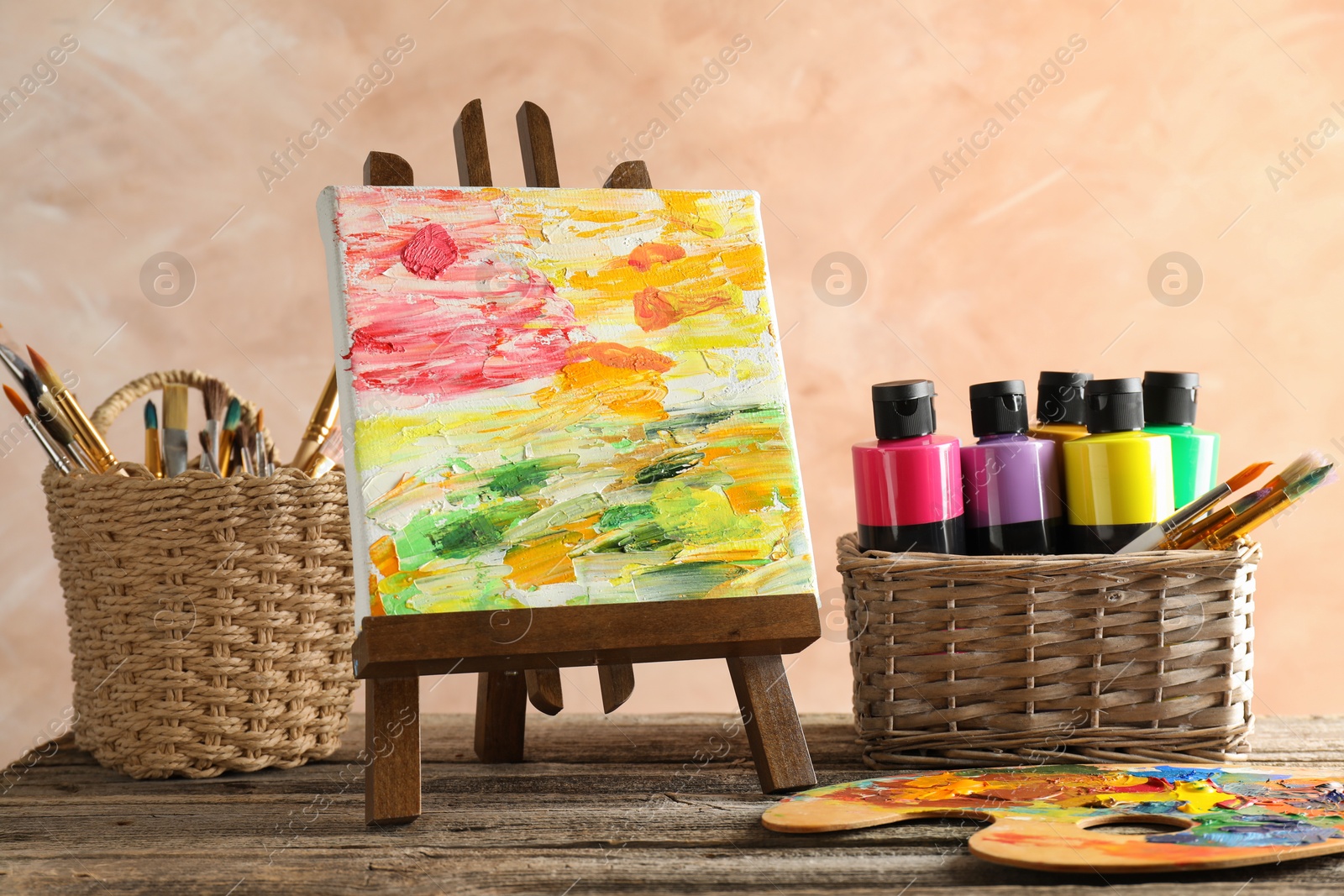 Photo of Acrylic paints of different colors, small easel with abstract picture, palette and brushes on wooden table