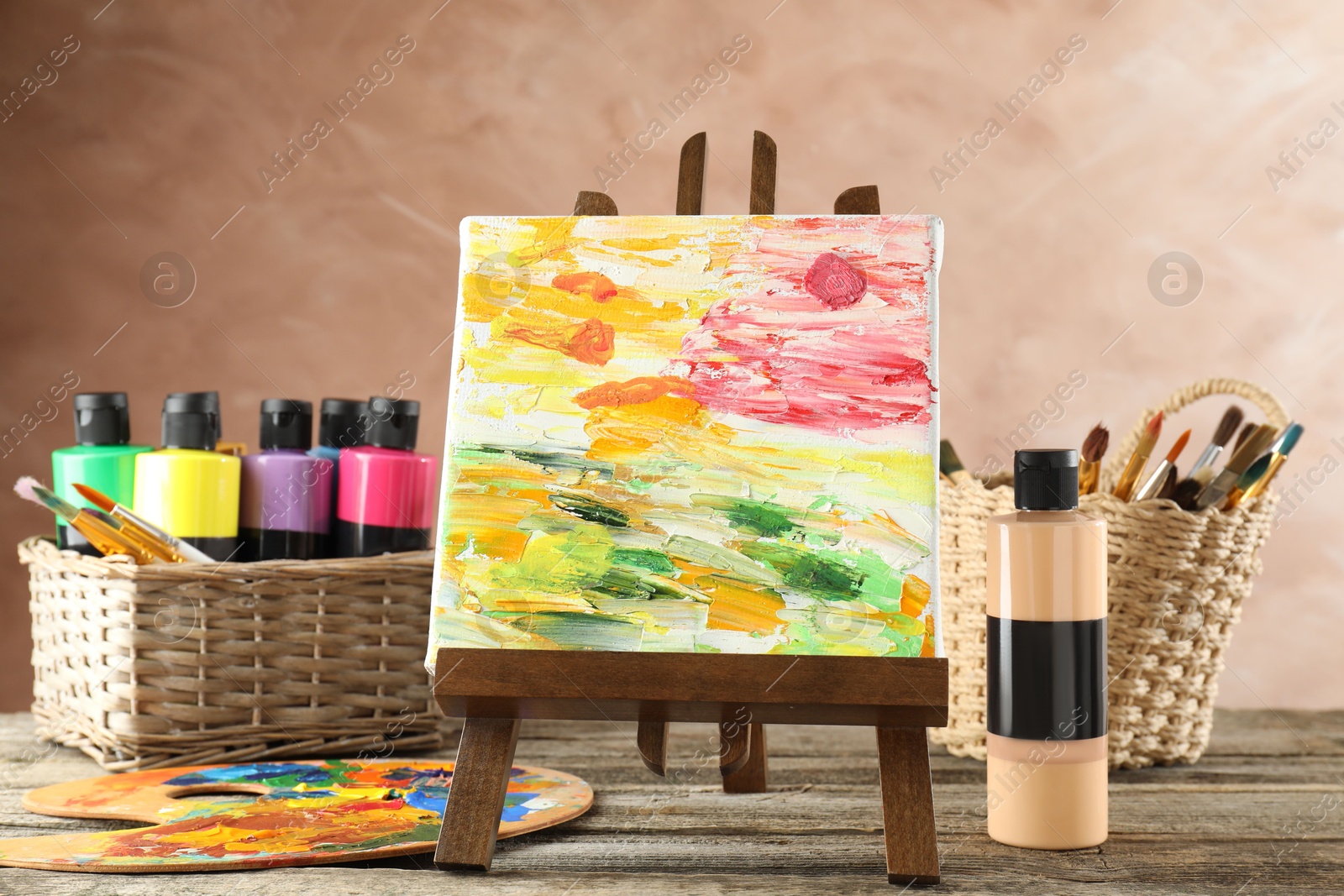 Photo of Acrylic paints of different colors, small easel with abstract picture, palette and brushes on wooden table