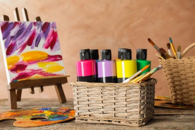 Photo of Acrylic paints of different colors, small easel with abstract picture, palette and brushes on wooden table